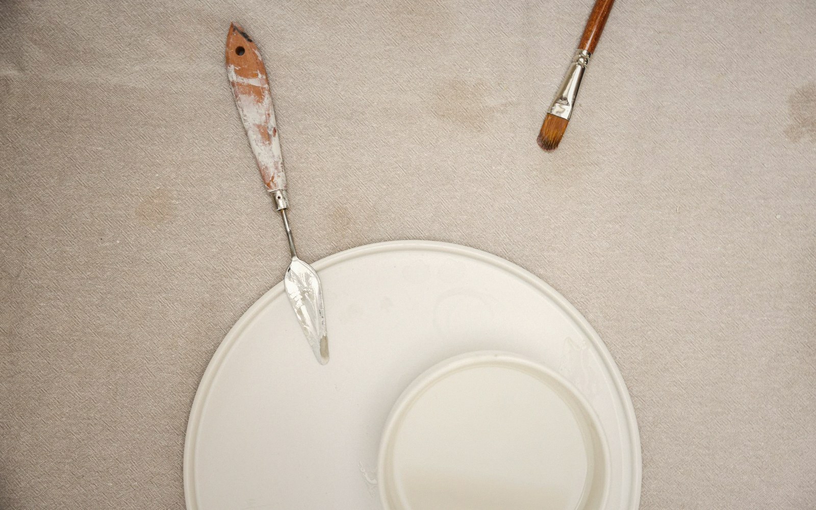 A wide flat brush loaded with warm white paint, tip pressed lightly against a primed canvas edge, palette in background
