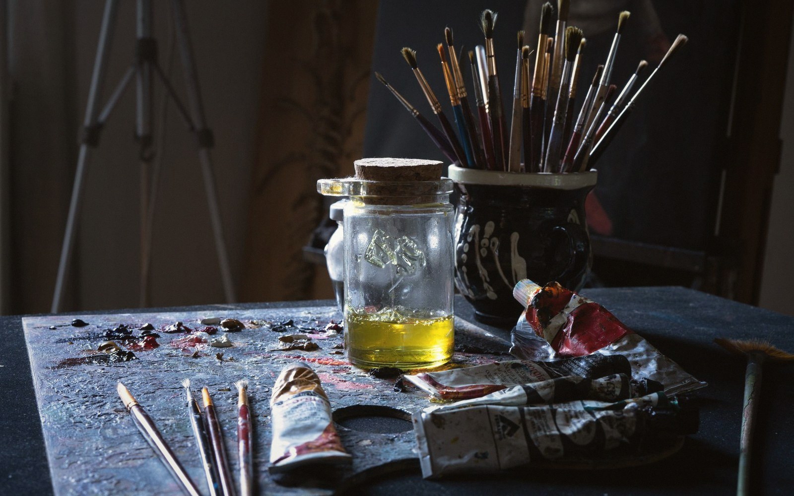 Paintbrushes stand in a jar beside a corked bottle of oil and paint tubes on a palette