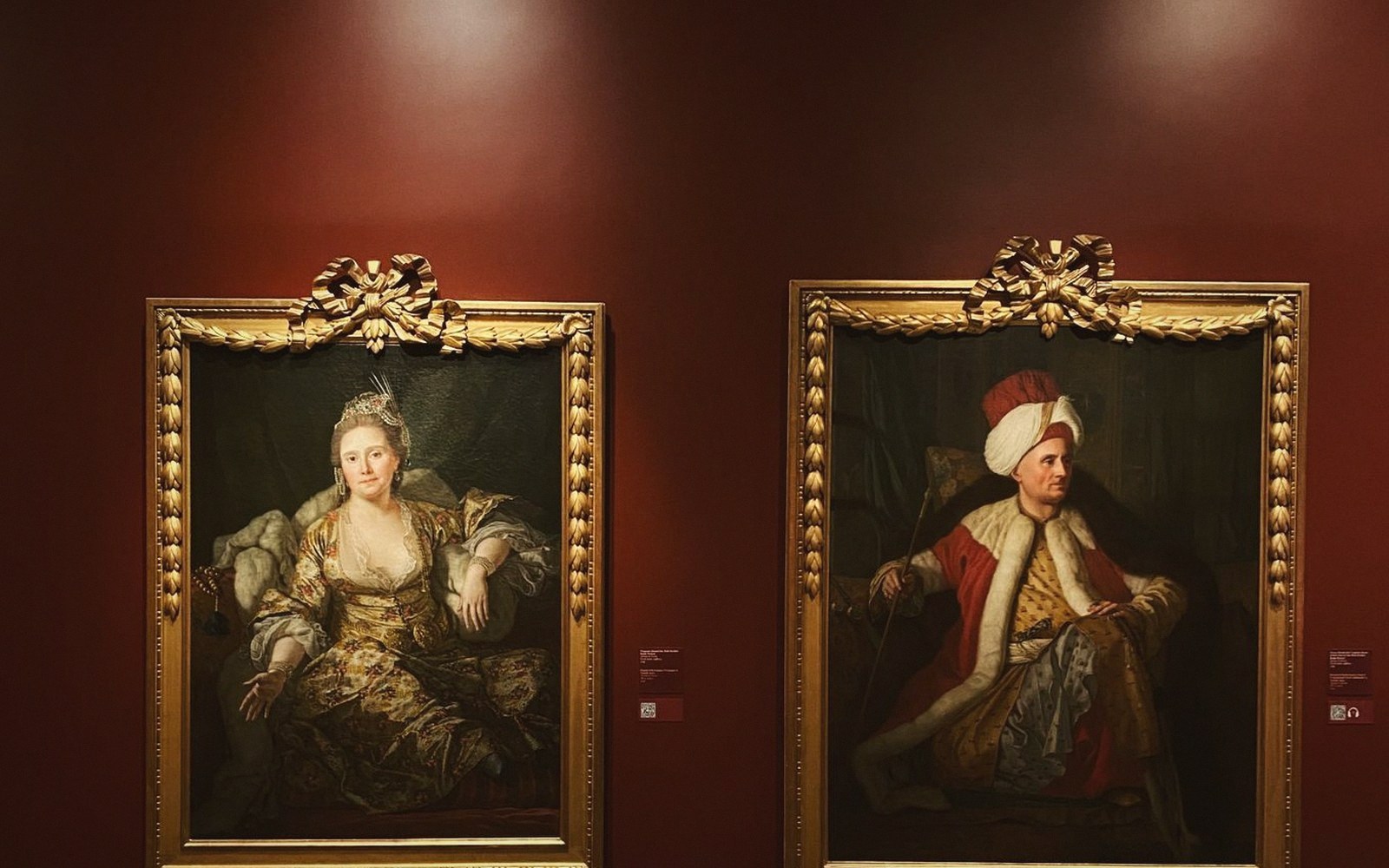 Two ornate gilt-framed portraits hang on a dark red gallery wall
