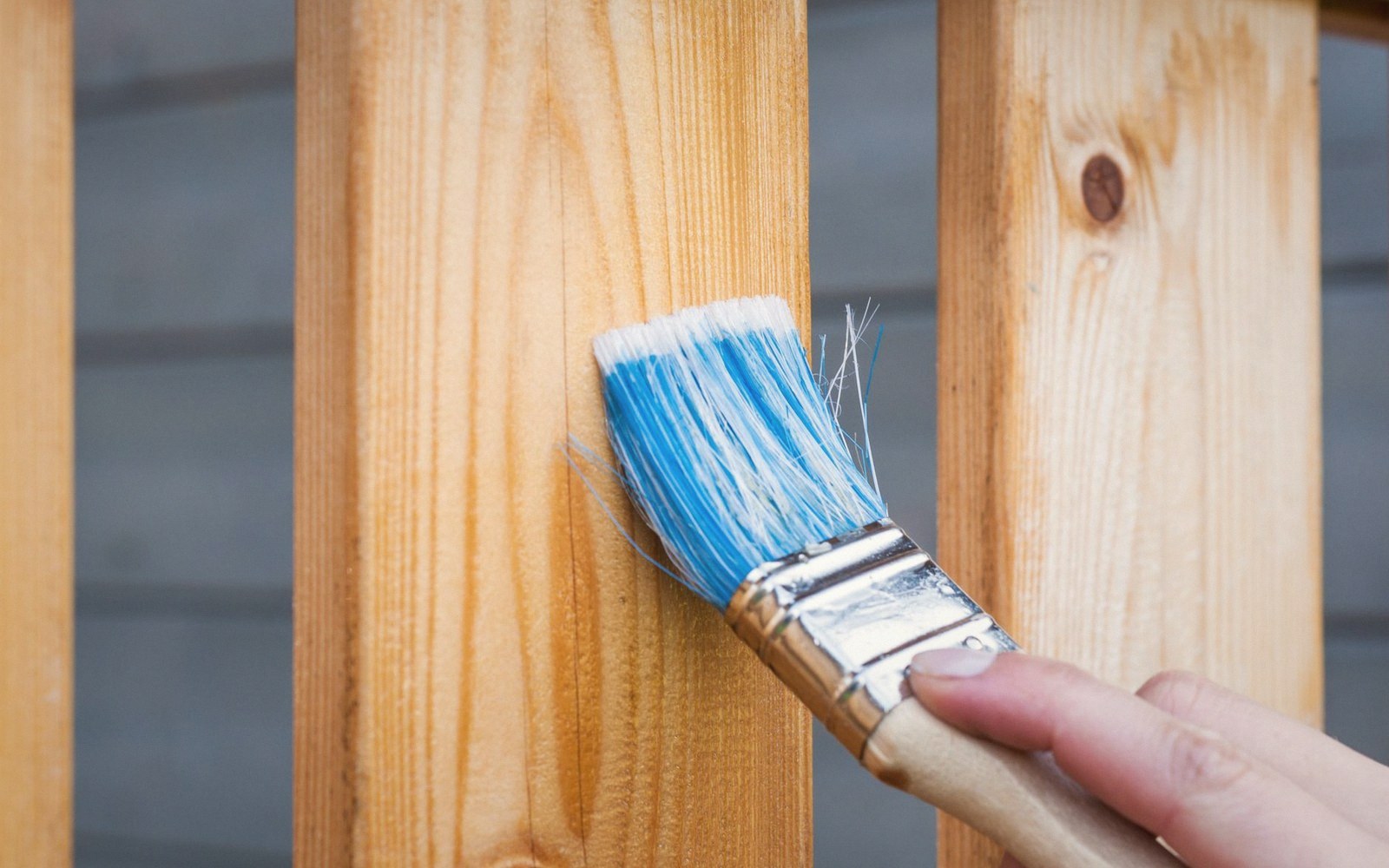 A hand uses a paintbrush to apply clear varnish to a wooden fence rail