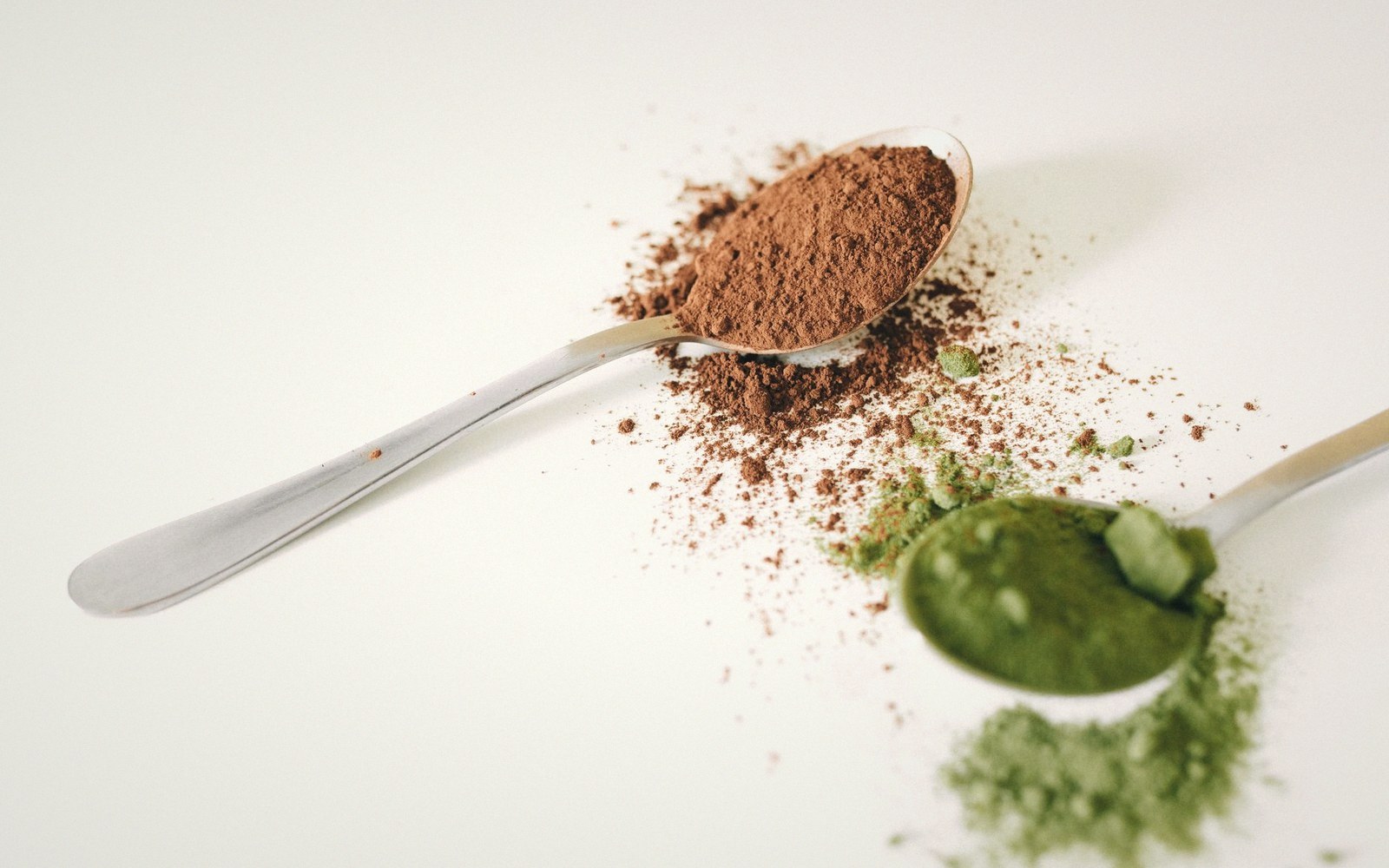Two spoons hold piles of cocoa powder and green matcha powder against a white background
