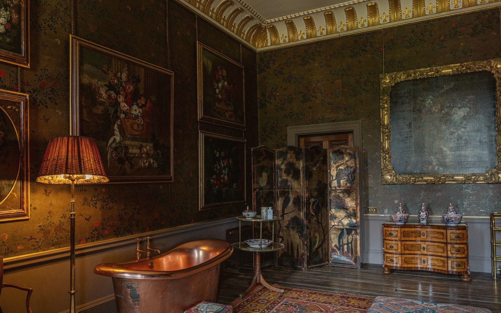 A copper bathtub sits beneath ornate gilded crown molding in a richly decorated period room