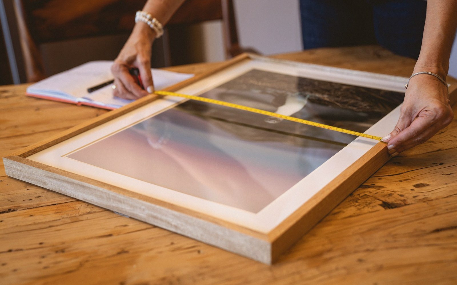 Hands measure a framed print with a tape measure while noting dimensions in a notebook