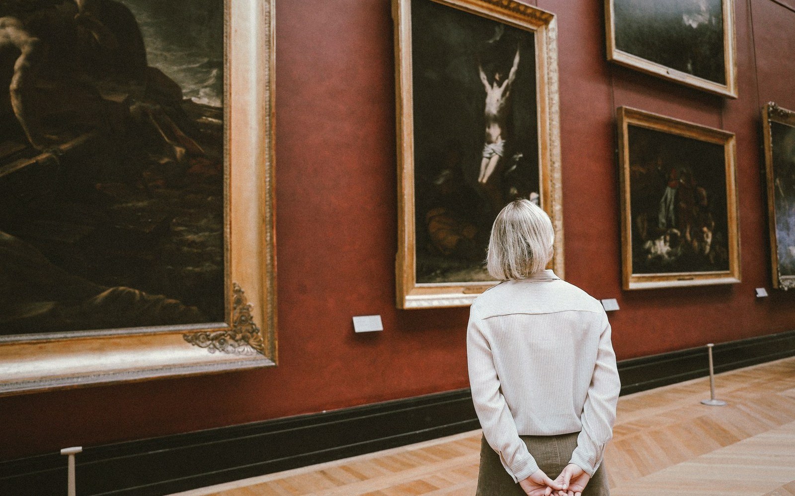 A woman views framed paintings on a red gallery wall lined with gold frames
