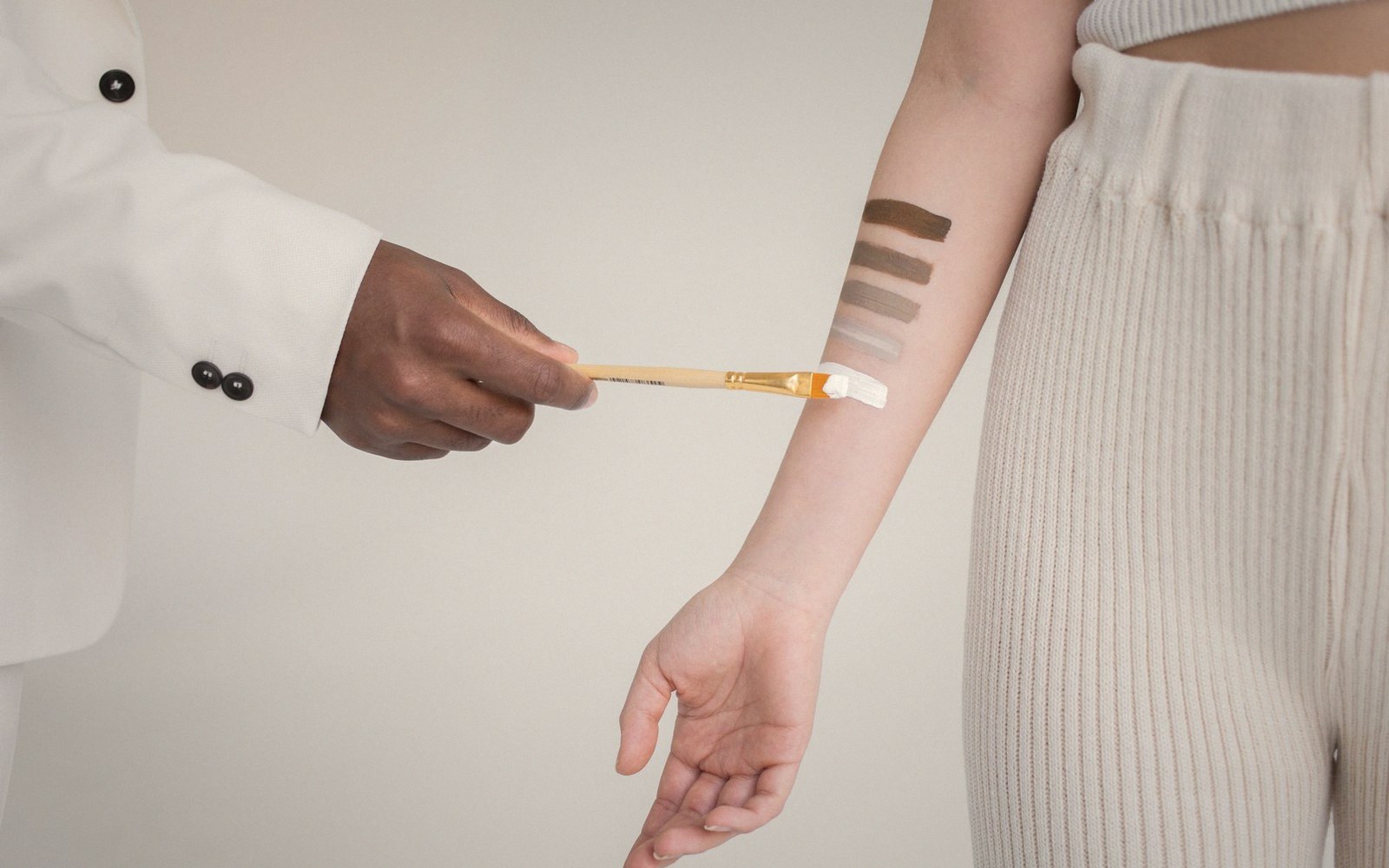 A hand uses a brush to swatch several shades of foundation on a forearm