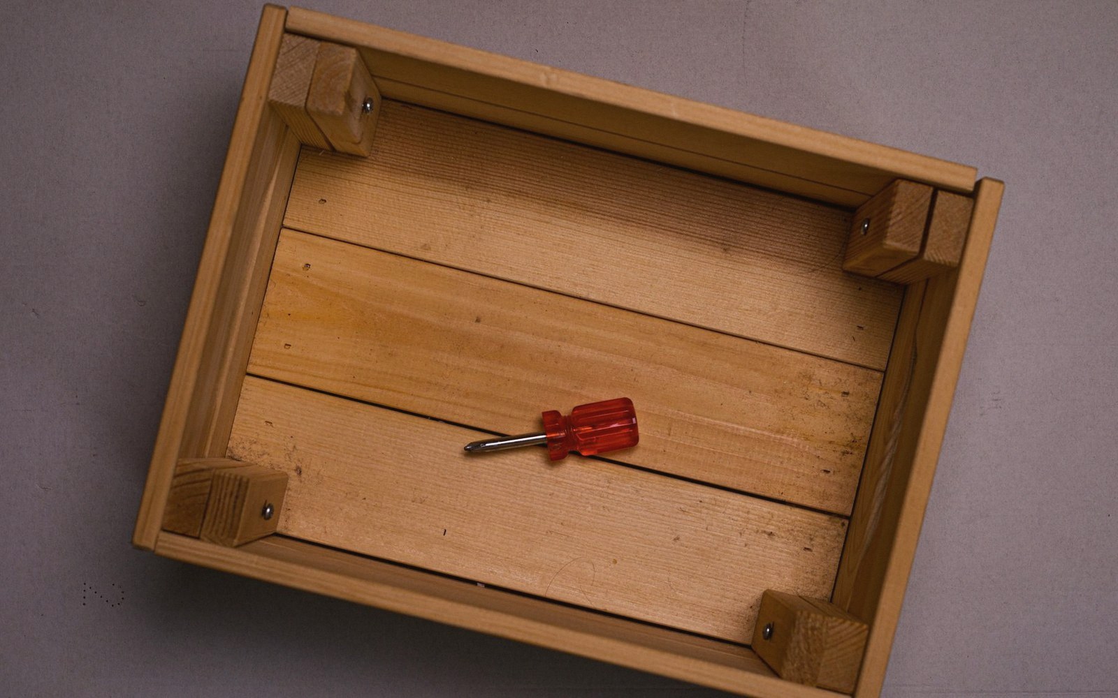 A red-handled screwdriver rests inside an empty wooden crate with corner blocks
